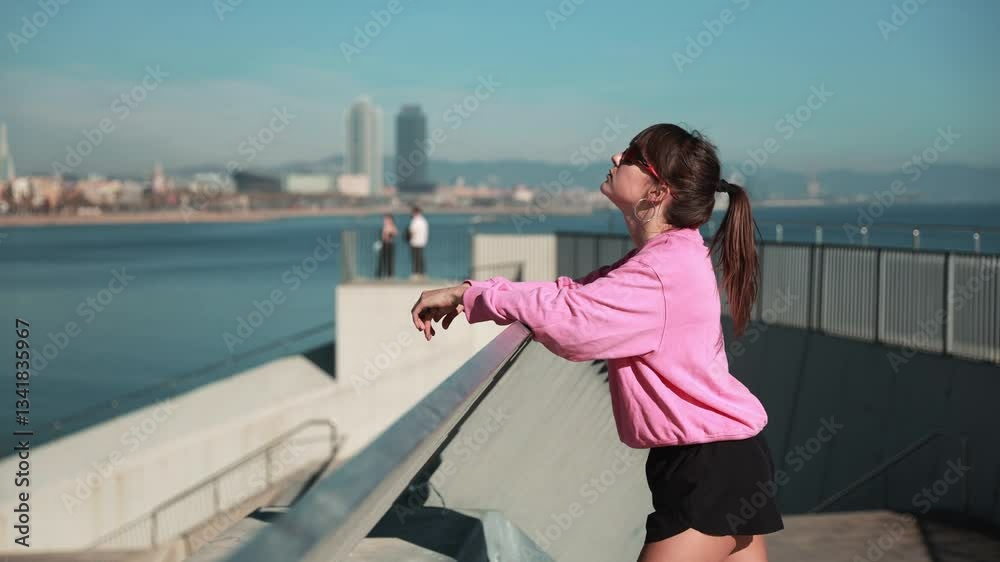 A young White woman in sunglasses and pink hoodie stops and takes a deep breath while walking outdoors along a waterfront promenade with modern city buildings and the ocean on a sunny day.