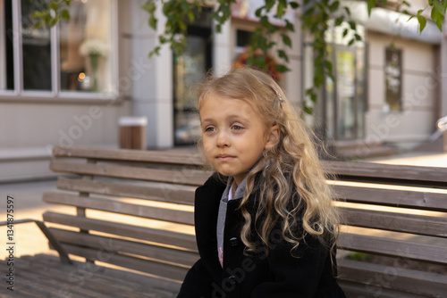 Young girl with long, curly blonde hair sitting alone on a bench on the street. The girl's expression is sad, unhappy, unsatisfied, she puckered her mouth,  A sad child left alone in a city