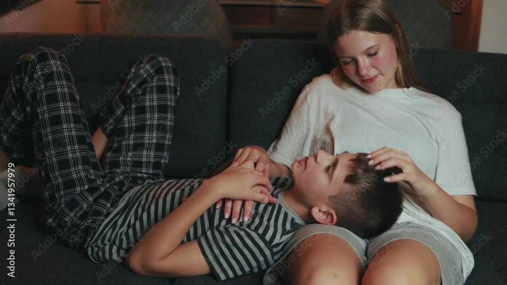 Teenage sister comforts her younger brother, who is lying on her lap on a comfortable sofa, creating a heartwarming scene of sibling affection
