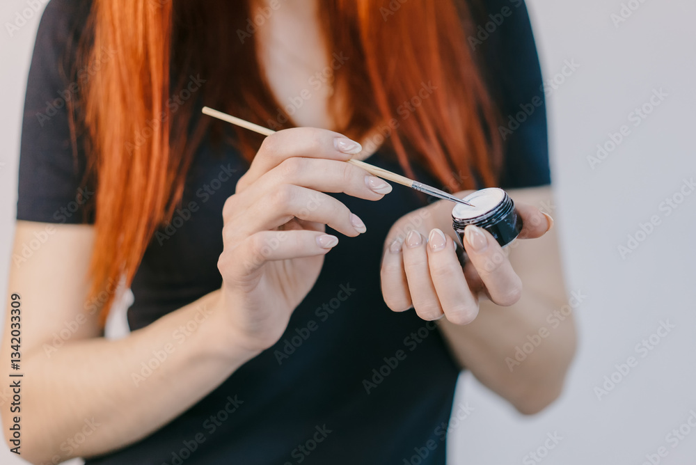 Fototapeta premium Applying makeup with precision brush. A person with long red hair applies makeup using a brush to a small container, demonstrating careful technique.