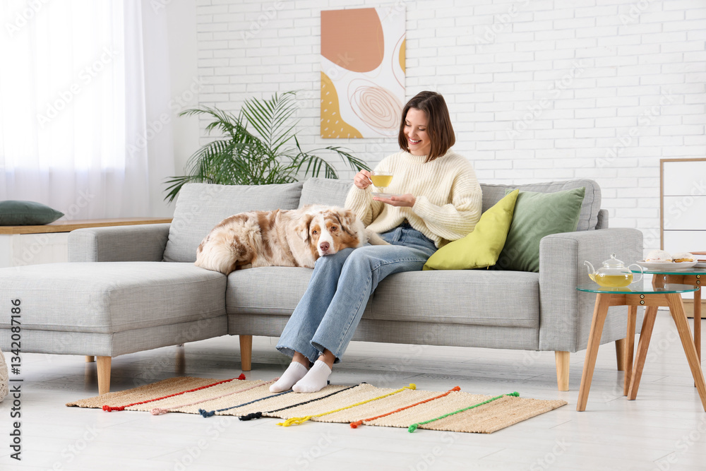 Fototapeta premium Beautiful young woman drinking green tea while sitting with her dog on sofa at home