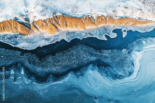 Aerial view of a frozen river flowing beside snow-covered, ochre-colored mountains.