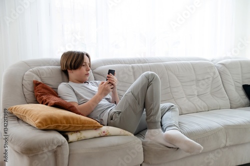 Teenager works on homework while relaxing on a couch in a cozy living room during the afternoon. Full length portrait of young teenage boy in hand smartphone while sitting on sofa