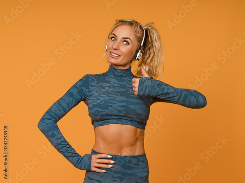 beautiful sports girl in sports camouflage clothes and headphones, smiling and showing thumb up on a yellow background, half-length portrait