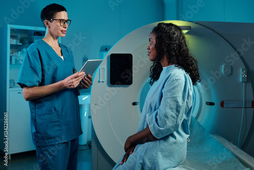 Medical professional discussing diagnosis with patient seated beside an imaging machine. Patient wearing hospital gown while doctor holds device, creating a focused environment in medical facility