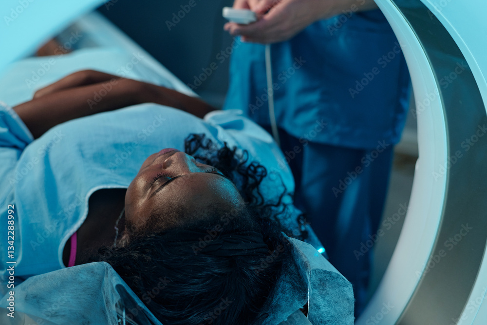 © pressmaster - African American patient at hospital lying in MRI scanner undergoing a medical procedure with nurse standing beside monitor in blue scrubs handling controls