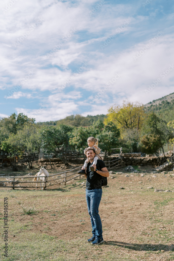 Dad with a little girl on his shoulders stands near a paddock with a horse in the park