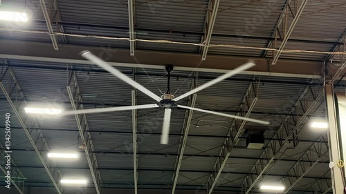 A large, industrial ceiling fan with five white blades spins in a high-ceilinged space, featuring exposed metal beams and rectangular light panels