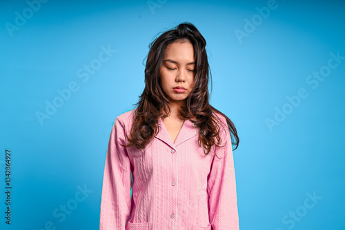 Asian woman in pink pajamas with messy hair and a sleepy, expressionless face, standing against a solid blue background, representing early morning mood or lack of sleep and energy