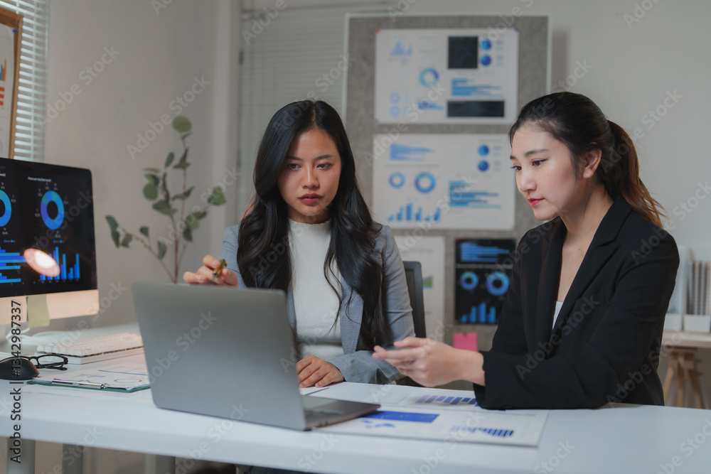 Two asian businesswomen analyzing charts and graphs on a laptop and printed documents during a productive meeting in a modern office, emphasizing collaboration and data-driven decisions Two asian businesswomen analyzing charts and graphs on a laptop and printed documents during a productive meeting in a modern office, emphasizing collaboration and data-driven decisions