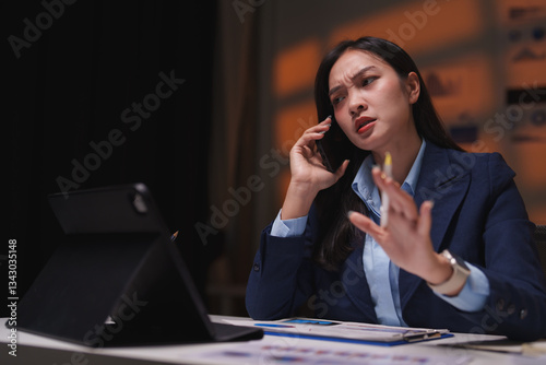 Young professional having a heated discussion on her mobile phone while reviewing documents on her tablet, working late into the night at a modern office