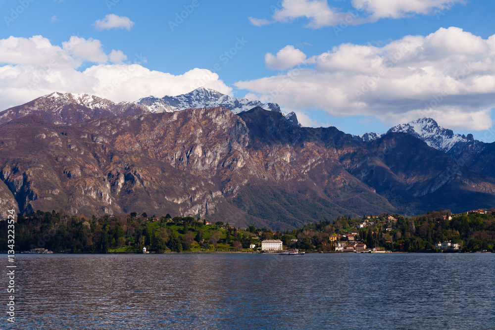 Obraz premium Wide panoramic scene of mountains and Como lake with hillside village nestled at the foot of the hills. Clear blue sky with scattered clouds add to peaceful and scenic atmosphere, Italy
