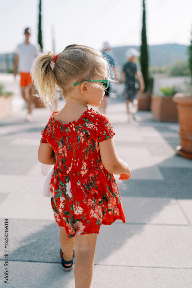 Little girl walks along the embankment with a pink toy and looks to the side. Back view Little girl walks along the embankment with a pink toy and looks to the side. Back view