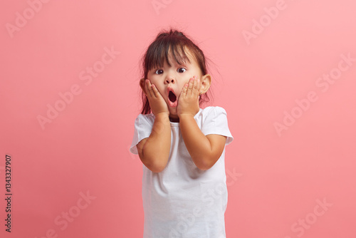 Amazed horrified little three years old girl keeps mouth opened, hands on cheeks, being shocked stares at camera, isolated over pink background. Kid expresses great surprisement. photo