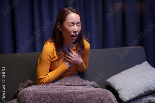 Young asian woman coughing and clutching her chest while sitting on a sofa at night, enduring the discomfort of a bad cold or flu, experiencing symptoms of illness and fatigue