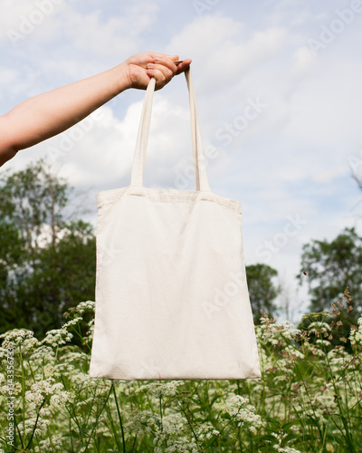 Mature senior woman, female hand holding white eco bag canvas fabric for mock up blank template on meadow with flowers background. Ecology, reusable concept. Eco friendly tote shopper