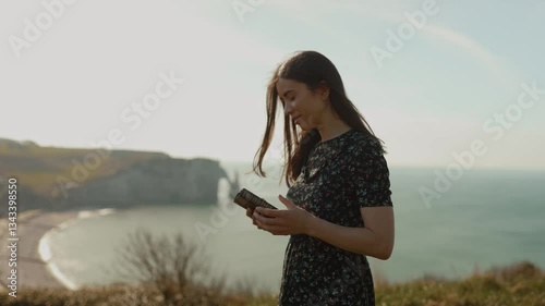 Portrait of a young woman girl holding a Bible in her hands, looking at the camera outdoors, calming smile on a rock in Etretat on the ocean shore during a trip, with a beautiful view in summer