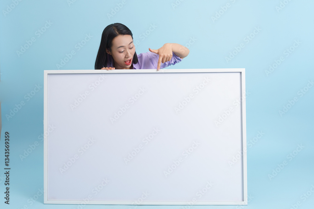 Excited young woman points at blank white canvas against turquoise background in creative studio setting Excited young woman points at blank white canvas against turquoise background in creative studio setting