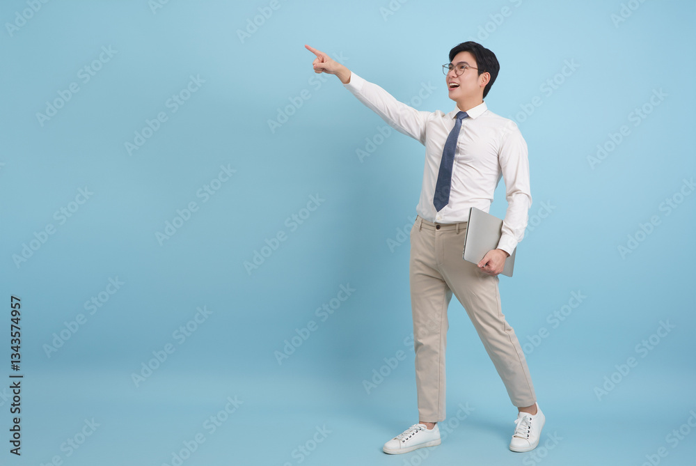 Young Asian professional in formal attire pointing towards a goal while holding a laptop against a light blue background Young Asian professional in formal attire pointing towards a goal while holding a laptop against a light blue background
