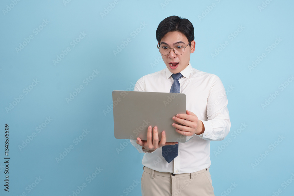 Excited young professional reacts to something surprising on laptop while standing against a blue background Excited young professional reacts to something surprising on laptop while standing against a blue background