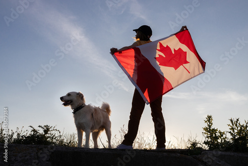 White terrier and woman holding large Canadian flag behind her against sunset sky. Walking with beloved pet. Happy Canada day. Freedom, trust, pride. Protect animals