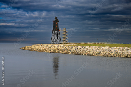 Ein einsamer Leuchtturm steht an einer felsigen Küste unter einem dramatisch bewölkten Himmel, der sich in ruhigen Gewässern spiegelt.