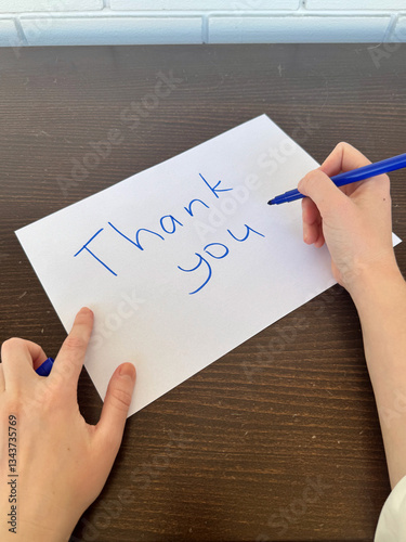 A close-up view of a white sheet of paper with the handwritten words Thank You placed on a dark wooden table, with two hands are nearby, one holding a blue marker