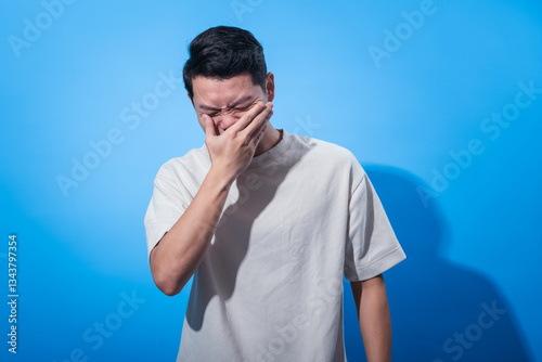 Asian man in a white t-shirt covering his nose and mouth with his hand while squinting his eyes tightly, expressing intense disgust or bad smell, standing against a bright blue background