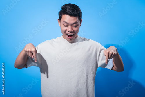 Excited young Asian man in beige t-shirt pointing downward with both hands while smiling and looking down against blue background, expressing surprise, direction, or attention in studio portrait