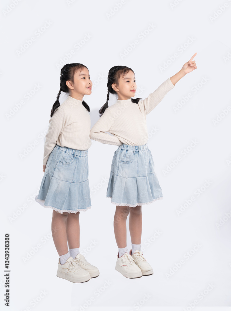 Naklejka premium Two young girls dressed alike, one pointing upwards, with similar outfits, on a clean, white background.