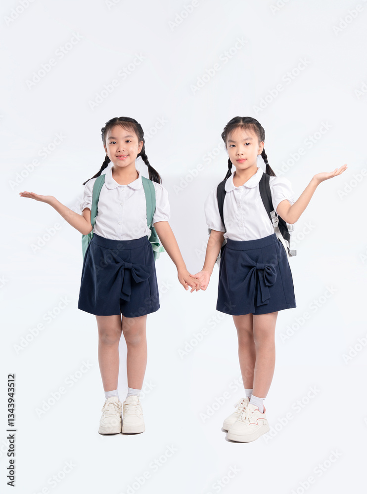Two young students, wearing school uniforms and backpacks, holding hands against a white backdrop. Two young students, wearing school uniforms and backpacks, holding hands against a white backdrop.