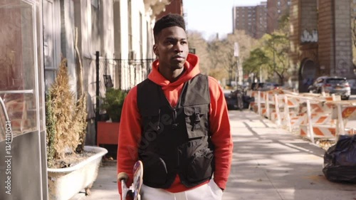 Young black male fashion model walking through the streets of New York City with Skateboard under his arm.
