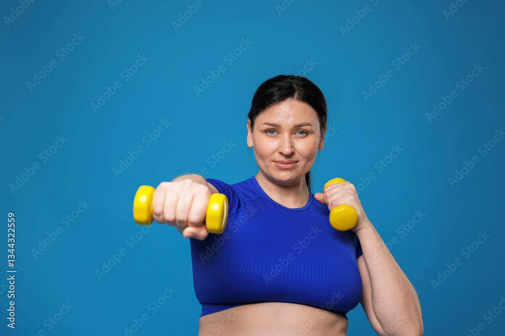 Plus size woman in gym clothes doing exercise with dumbbells on light blue background