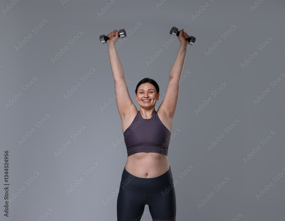 Plus size woman in gym clothes doing exercise with dumbbells on grey background