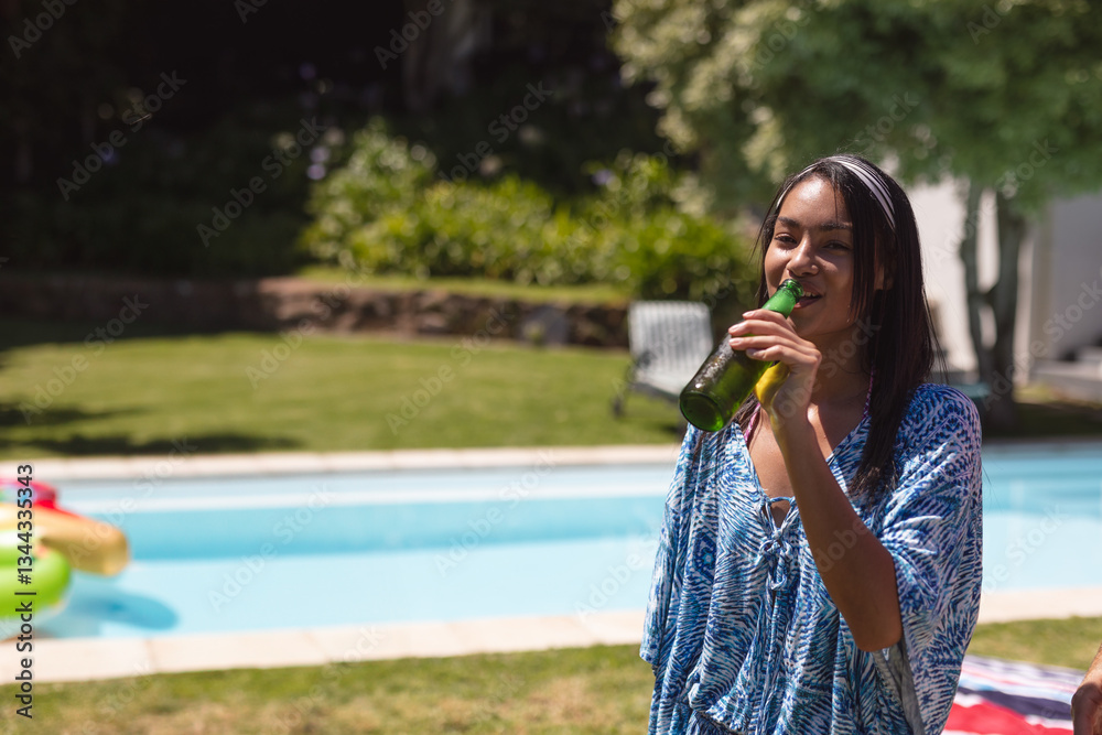 Fototapeta premium Young woman enjoying cold drink by pool, smiling and relaxing at outdoor party, copy space