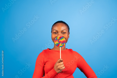 Studio portrait of a cheerful young woman holding a rainbow lollipop in front of her mouth, enjoying a sweet treat