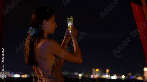 A young woman enjoys a magical night view from a mountain in an amusement park, recording the moment on her phone. A traveler stands on a mountain in an amusement park, taking a video.