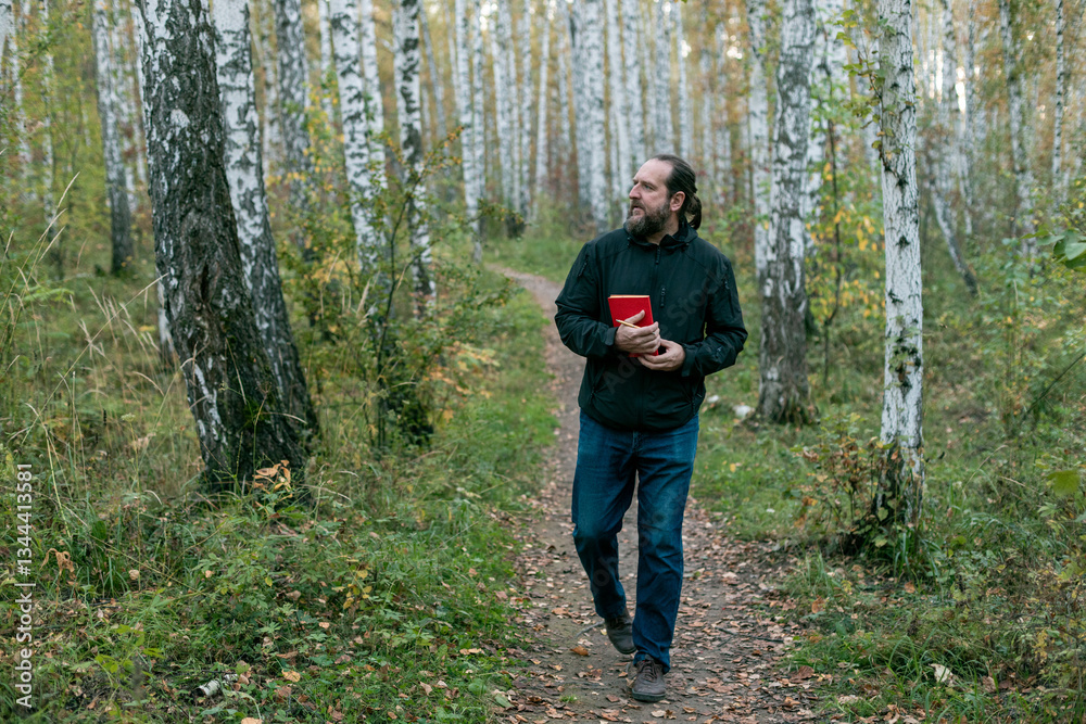 Fototapeta premium a bearded man walking through the forest with a red book and looking into the forest