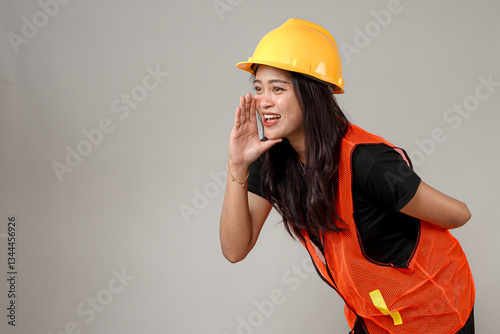 Smiling Asian woman in a yellow hard hat and orange safety vest shouts or calls out with an enthusiastic and confident expression. She appears energetic and expressive.