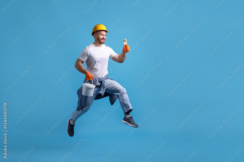 Man wearing hardhat with brush and bucket of paint jumping on blue background