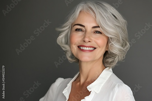 A radiant mature woman with a captivating smile, showcasing her elegant silver bob hairstyle against a simple gray backdrop.