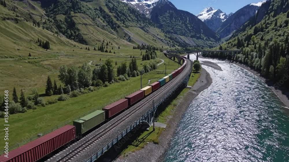 Freight train travels alongside a river in a picturesque mountain valley, surrounded by lush greenery and snow capped peaks under a clear blue sky