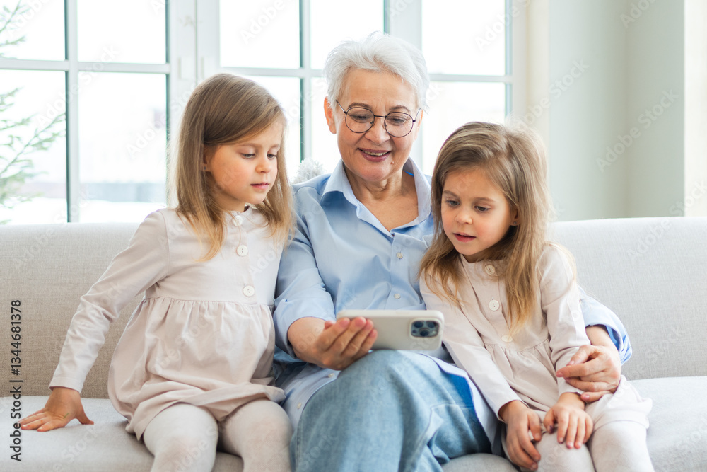 Obraz premium Happy family at home. Two little girls sisters twins grandmother enjoying time together watching video on phone. Good time at home. Grandma granddaughters child kids together. Family generations