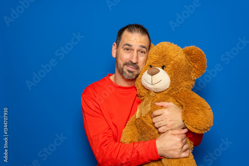 Bearded Hispanic man in his 40s wearing a red sweater energetically hugging his teddy bear, isolated on blue studio background.