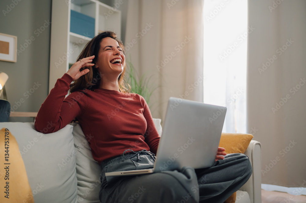 Fototapeta premium Happy woman laughing while using laptop on sofa at home