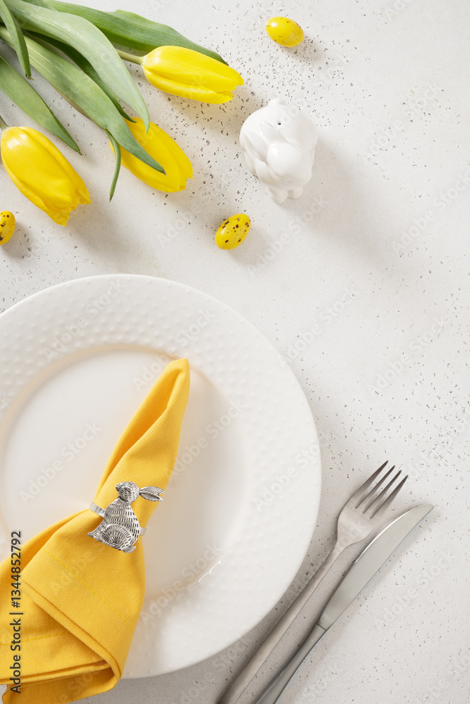 Easter table setting with spring, yellow tulips, white plates, bunny, eggs on white background. Happy Easter celebration. Copy space. Vertical. View from above.