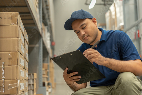 Warehouse worker wearing a hat and black shirt checking quality by clipboard on tall shelves in warehouse storage. Asian auditor or staff work looking up stock taking inventory in warehouse store.