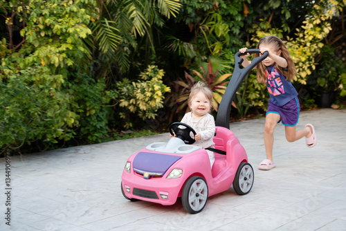Happy baby in a pink toy car smiles while gripping the steering wheel, as an older sister joyfully pushes from behind capturing sibling fun and outdoor playtime. Authentic, candid shots