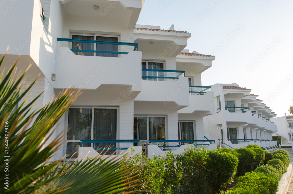 Obraz premium White resort exterior under bright sunlight features large balconies and lush green hedges. Modern architectural design with clean lines viewed at angle enhances its appeal