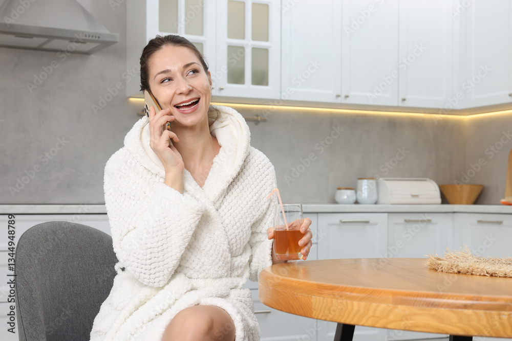 Beautiful woman with glass of juice talking on smartphone after spa procedure in kitchen Beautiful woman with glass of juice talking on smartphone after spa procedure in kitchen